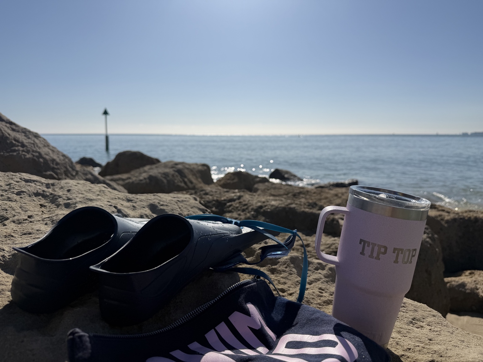 Swimming fins, goggles and a pink travel mug on sunlit rocks beside the sea